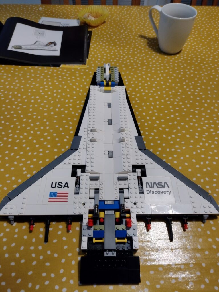 Space shuttle body covered in white flat panels, laid out vertically nose towards the top. The internal bricks that make up the structure are no longer exposed. 