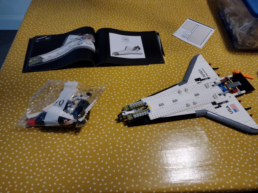 Flat space shuttle body laid flat. Next to the shuttle are bag 10 of bricks and the Lego manual showing the shuttle with the three main rocket boosters installed on a panel that juts inwards at a slight angle from vertical relative to the body.
